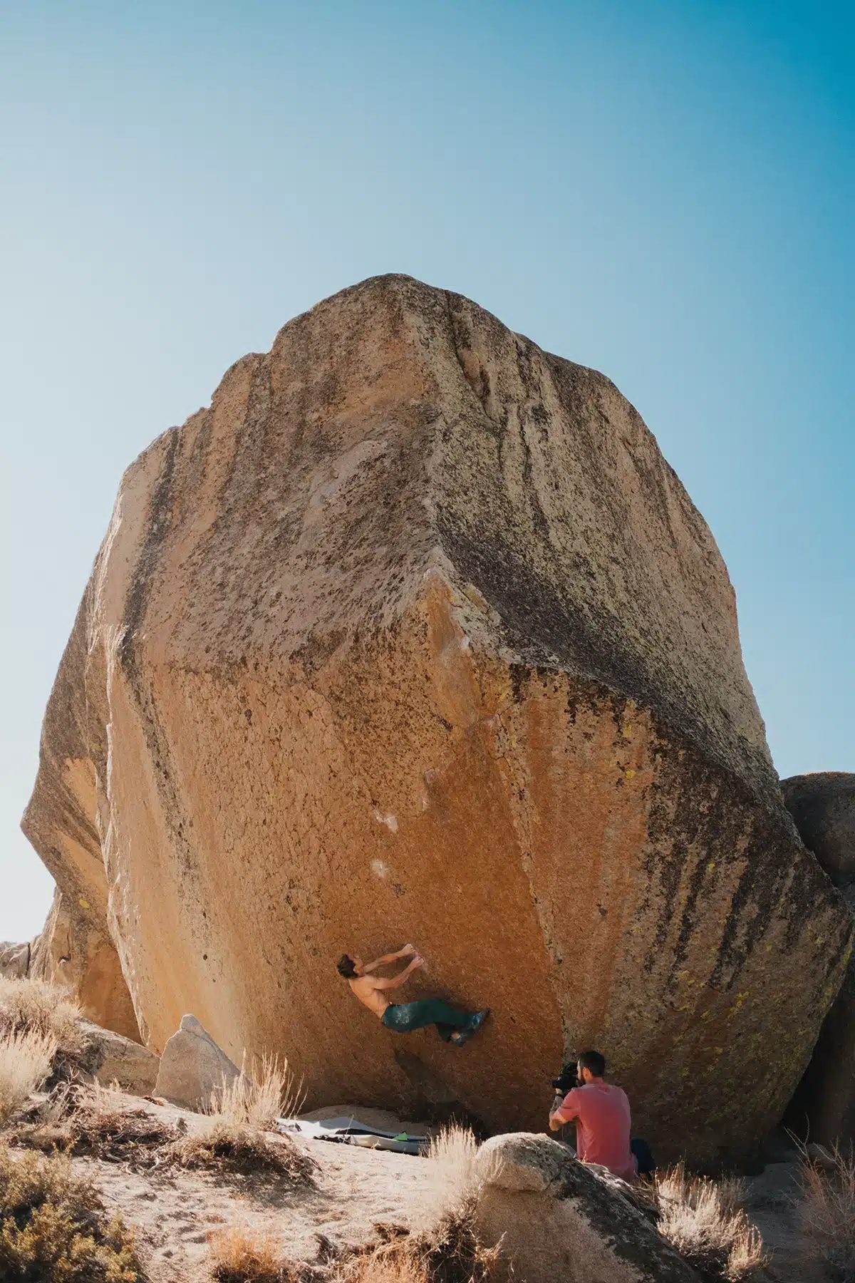 shirtless climber wearing teal pants is bouldering on a large, angular rock formation. The rock has a rough, textured surface with patches of moss. Below, a photographer in a pink shirt captures the scene, and crash pads are visible on the ground. The sky is clear and blue.