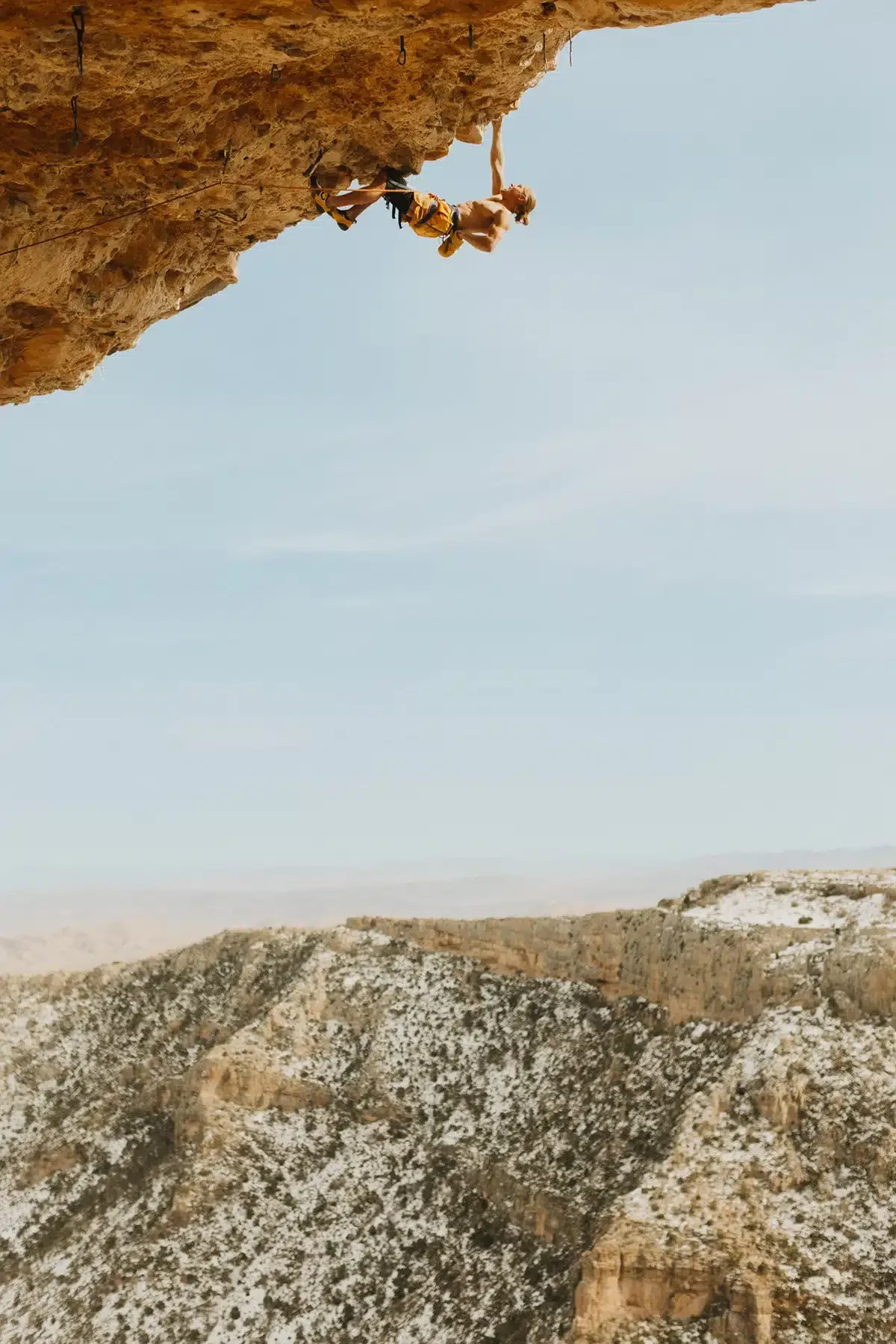 A shirtless rock climber wearing yellow shorts is hanging from an overhanging rock formation. He is gripping small holds with both hands, with a rope for safety. The background features a rugged landscape with mountains under a clear blue sky.