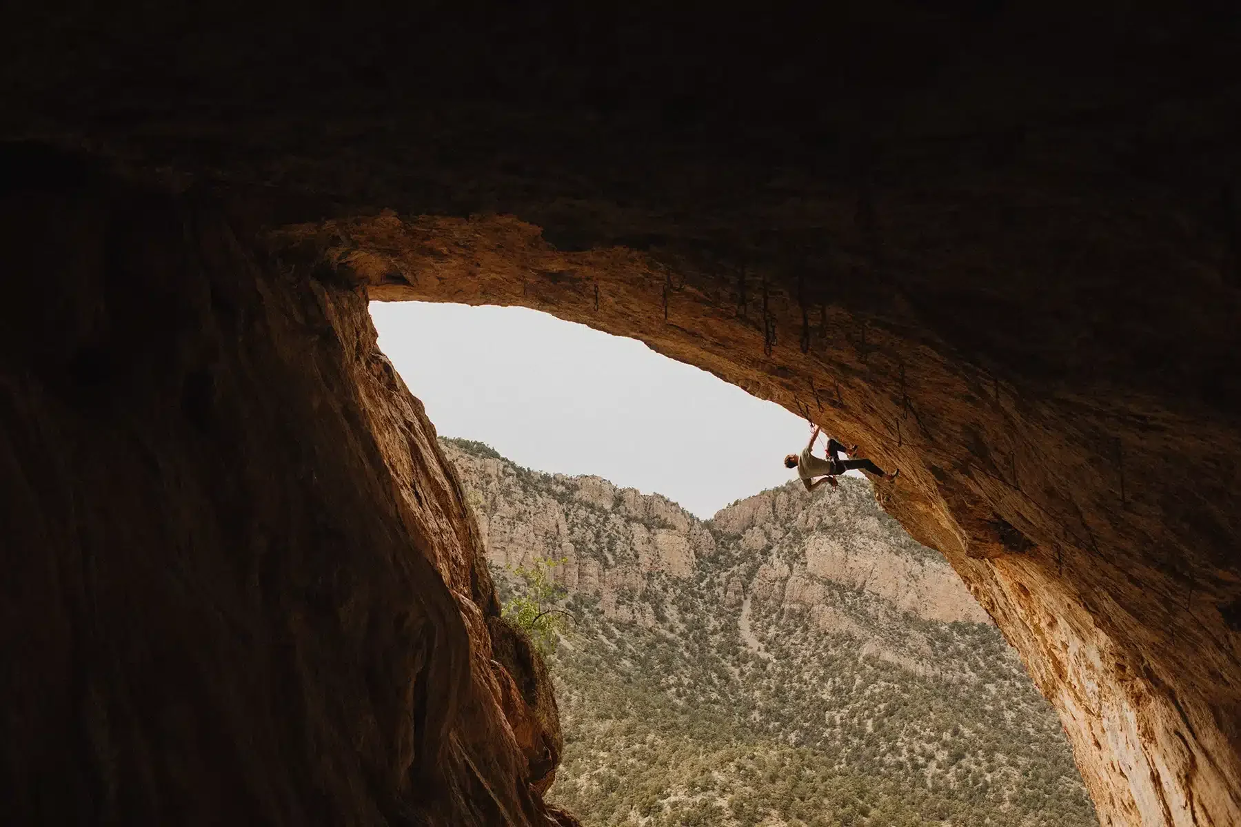 A rock climber is hanging from the roof of a large cave with rugged, reddish rock formations. The climber is secured by a harness and rope, and a mountain range covered in green trees is visible in the background through the cave opening.