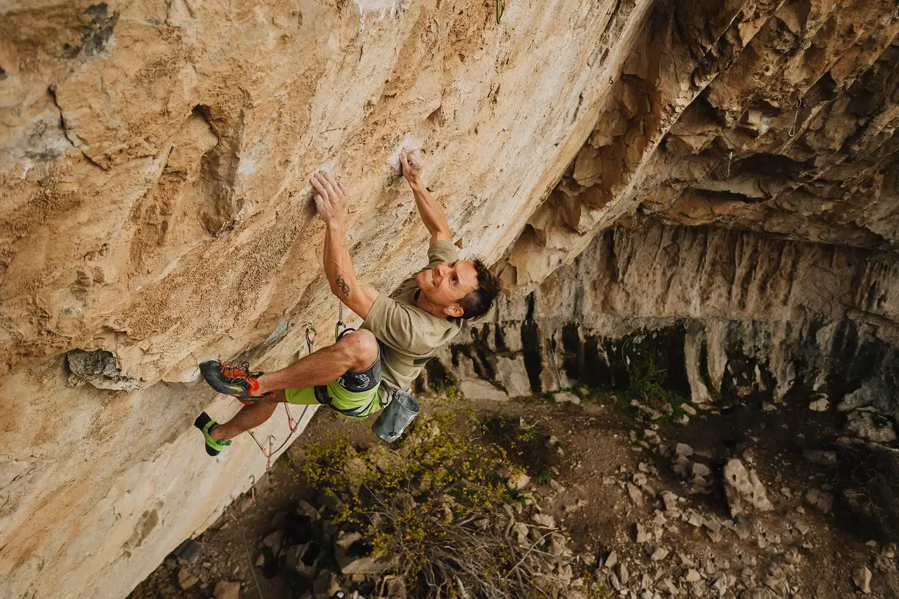 A rock climber in a beige shirt and green shorts is gripping a vertical rock wall. His face shows determination as he carefully navigates the rough, tan-colored rock. The ground below is scattered with rocks and sparse vegetation.