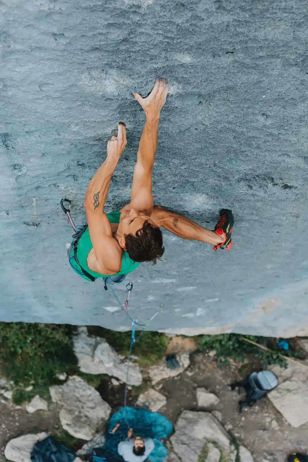 A rock climber wearing a green tank top and red climbing shoes is scaling a steep grey rock face. His arms are extended as he reaches for small holds. The view is from above, showing the climber's intense focus and the belayer below, standing on rocky ground with climbing gear.