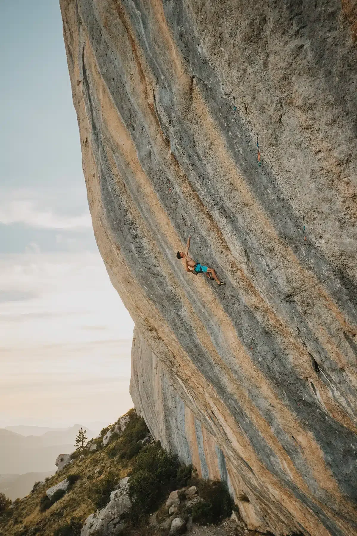 A shirtless rock climber wearing blue shorts is scaling a steep, striped rock face. The rock has distinct vertical bands of grey and tan. The landscape below shows a rugged terrain with bushes and a few scattered rocks, set against a backdrop of distant mountains under a clear sky.