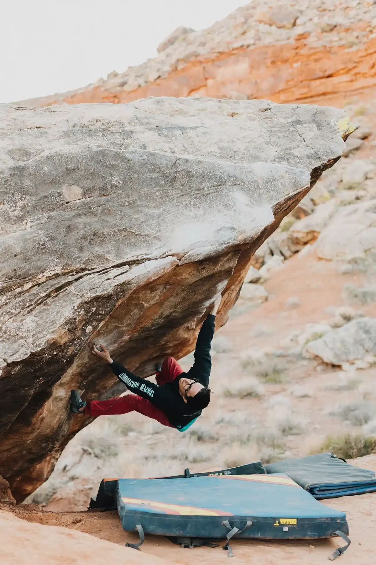 A climber wearing a black shirt and red pants is bouldering on a large, overhanging rock formation. His right hand grips a small hold while his left hand reaches up. The ground below has crash pads for safety, and the background features reddish rock and sparse desert vegetation.