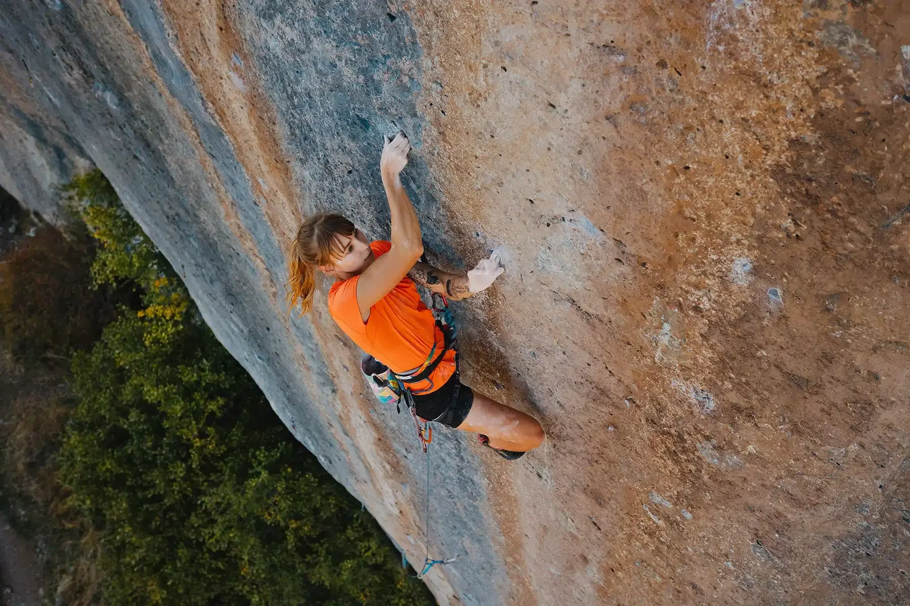 A woman wearing an orange top and black shorts is climbing a steep rock face. Her left hand is reaching for a small hold while her right hand grips the rock. The view is from above, showing the expanse of the rock and the greenery below.