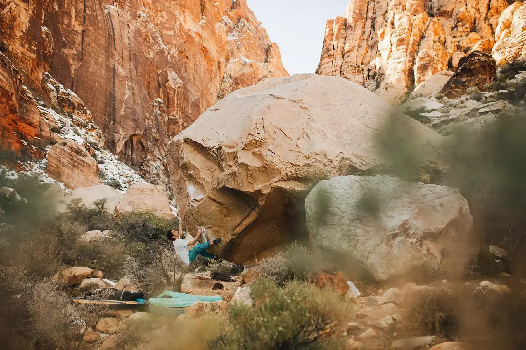 A rock climber wearing a white shirt and blue pants is bouldering on a large, overhanging boulder in a canyon with reddish rock walls. The scene includes climbing pads at the base of the boulder and sparse vegetation around.